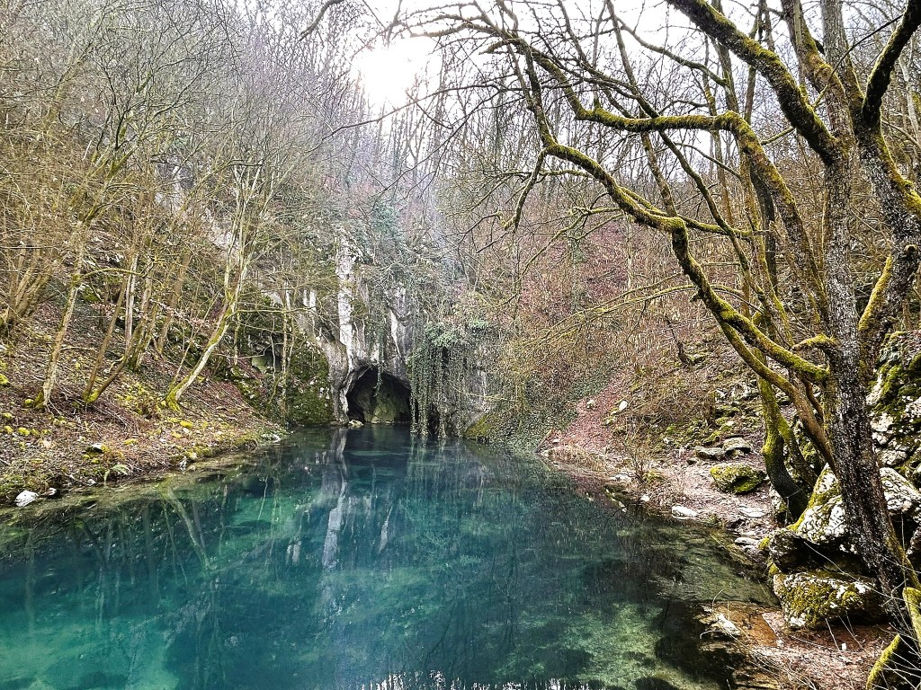A serene natural scene featuring a still, clear blue pond surrounded by bare trees and rocky terrain, with a cave entrance visible in the background.