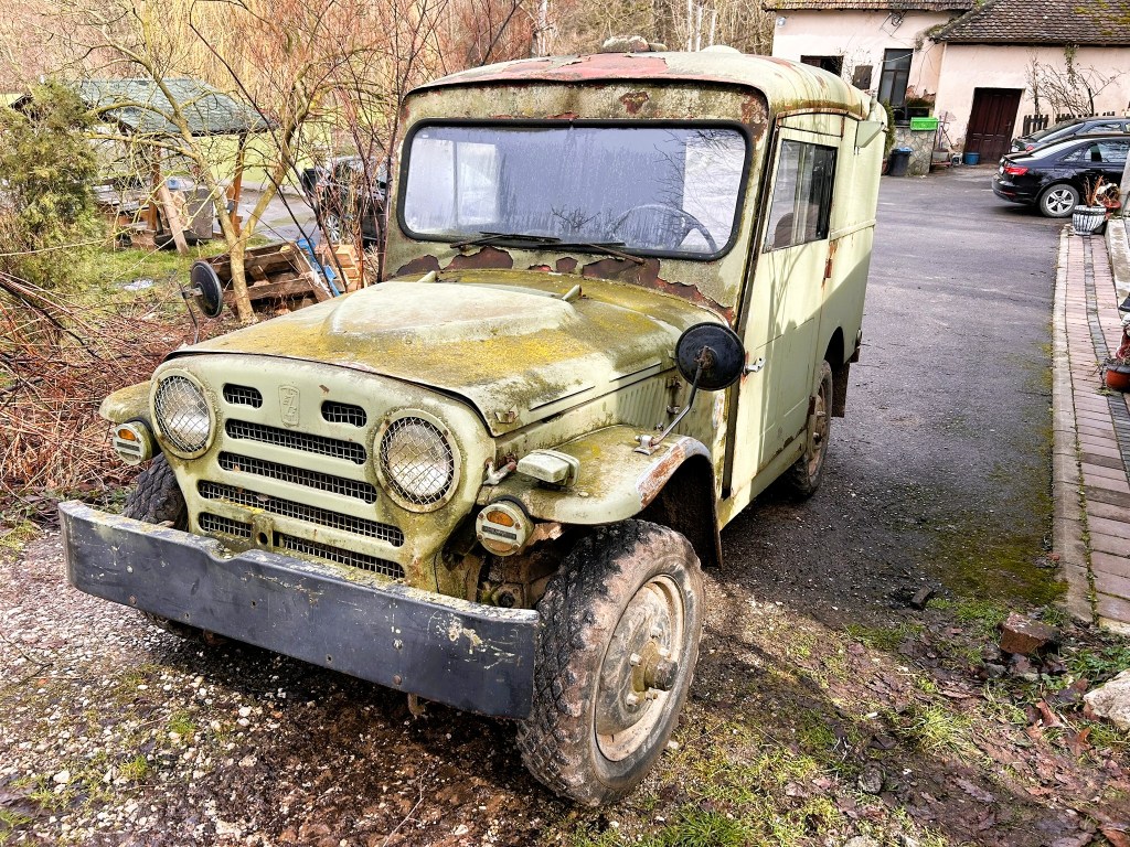 An old, moss-covered green vehicle parked on a gravel path, showing signs of rust and deterioration, with a house and cars visible in the background.