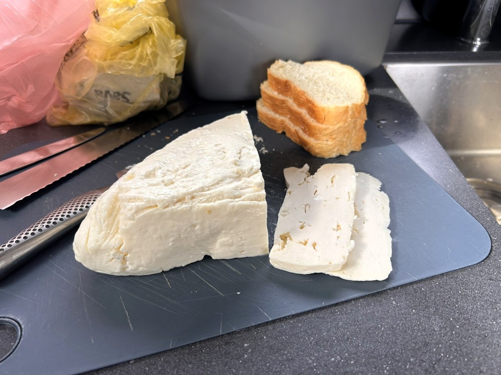 A cutting board with a block of cheese and sliced cheese, accompanied by several slices of bread and utensils.