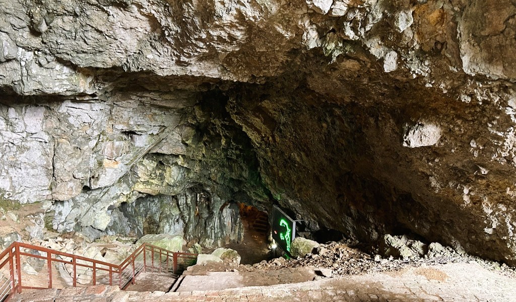 An interior view of a cave entrance with rocky walls and a staircase leading down into the cavern. Soft lighting and a green sign are visible in the background.