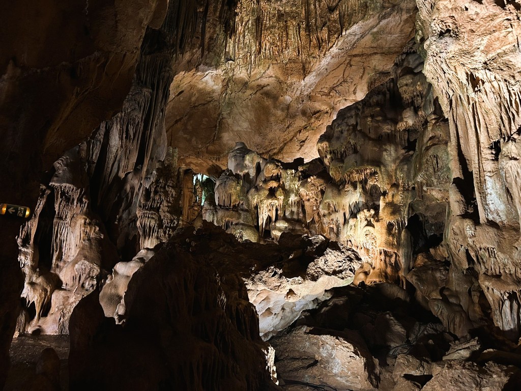 A large underground cave with impressive stalactites and stalagmites, illuminated by soft lighting.