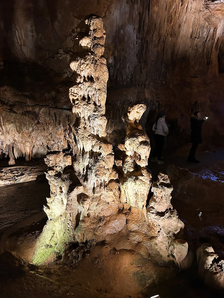 A tall, textured rock formation illuminated in a cave setting, with a few visitors exploring the area in the background.