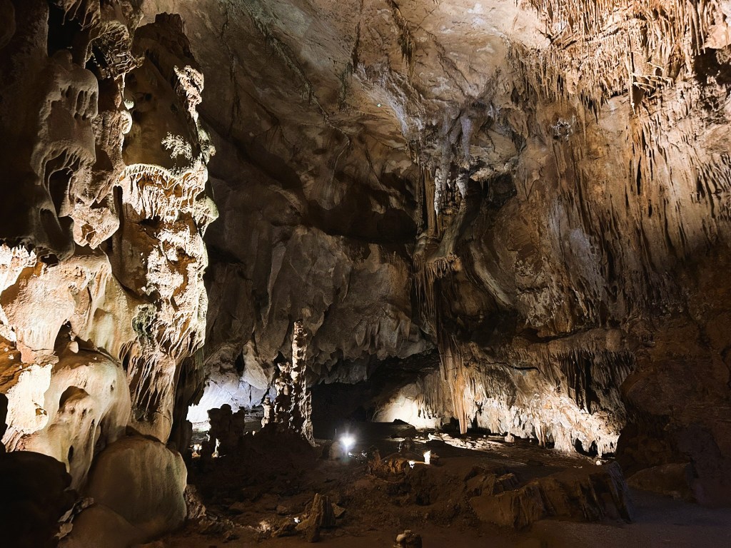 An illuminated cave interior featuring stalactites and stalagmites, with rocky walls and various textures.