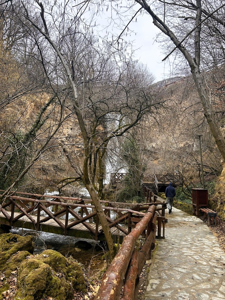 A man walking along a stone pathway beside a serene stream, with wooden bridges and bare trees in a winter landscape.