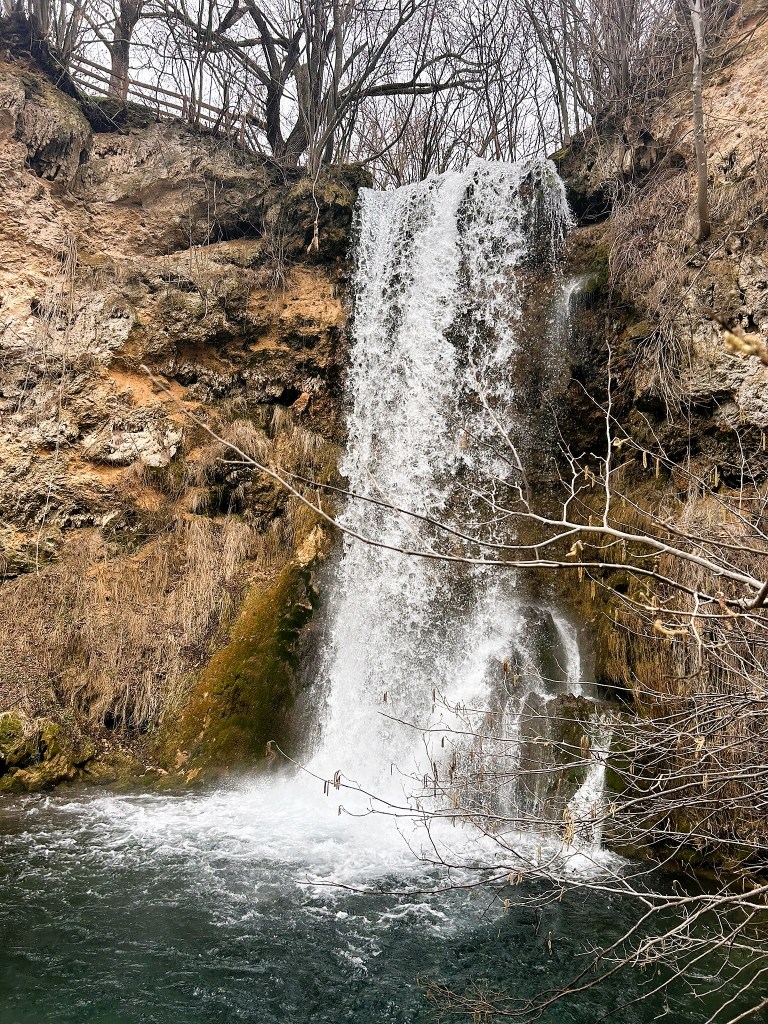 A waterfall cascading down a rocky cliff surrounded by bare trees and dry vegetation, with water splashing into a pool below.