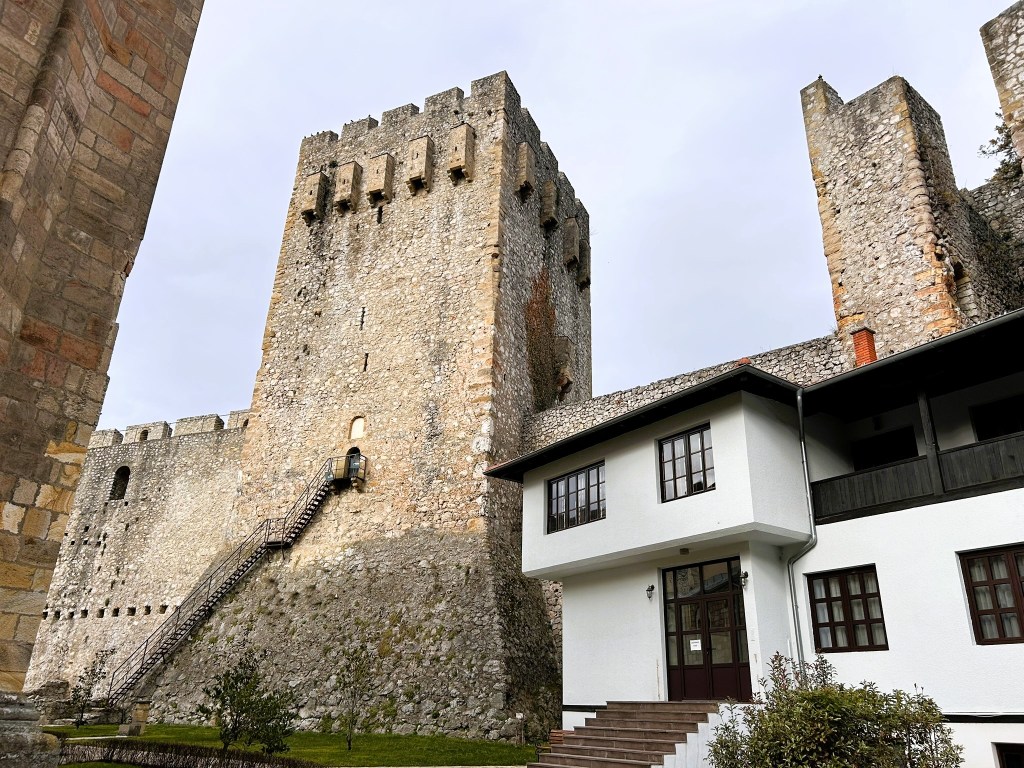 View of a historic stone castle tower with a wooden staircase leading to its entrance, alongside a modern white building and remnants of castle walls.