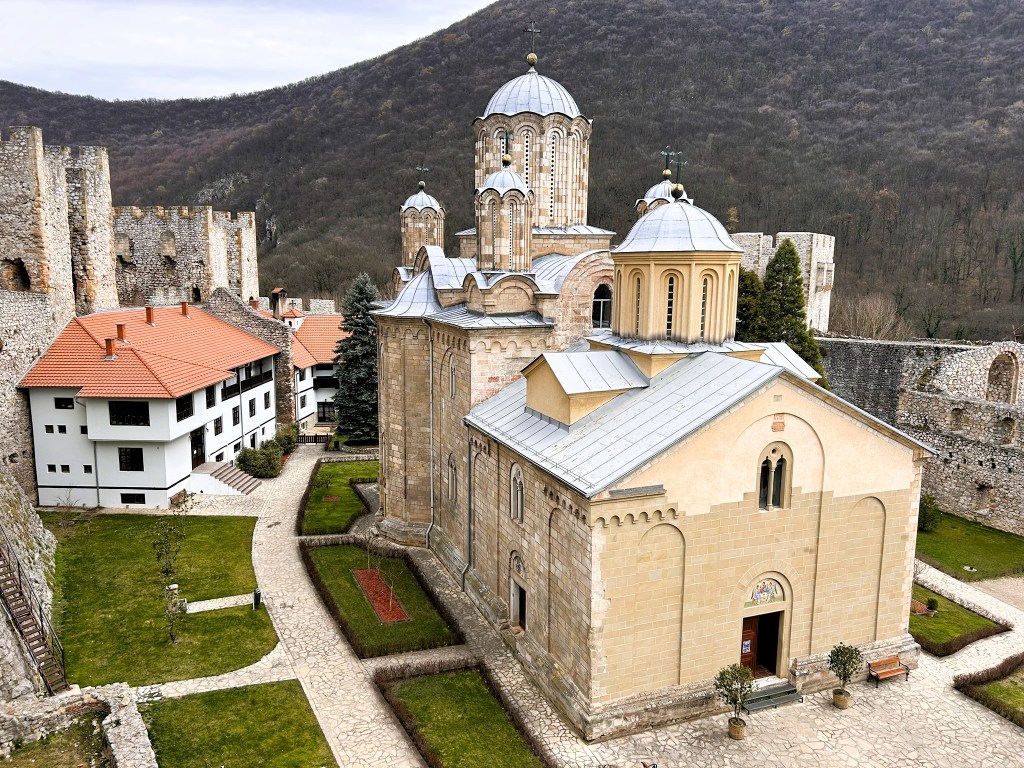 Aerial view of a historic monastery complex with stone walls, multi-domed church, and adjacent residential buildings, set against a mountainous backdrop.
