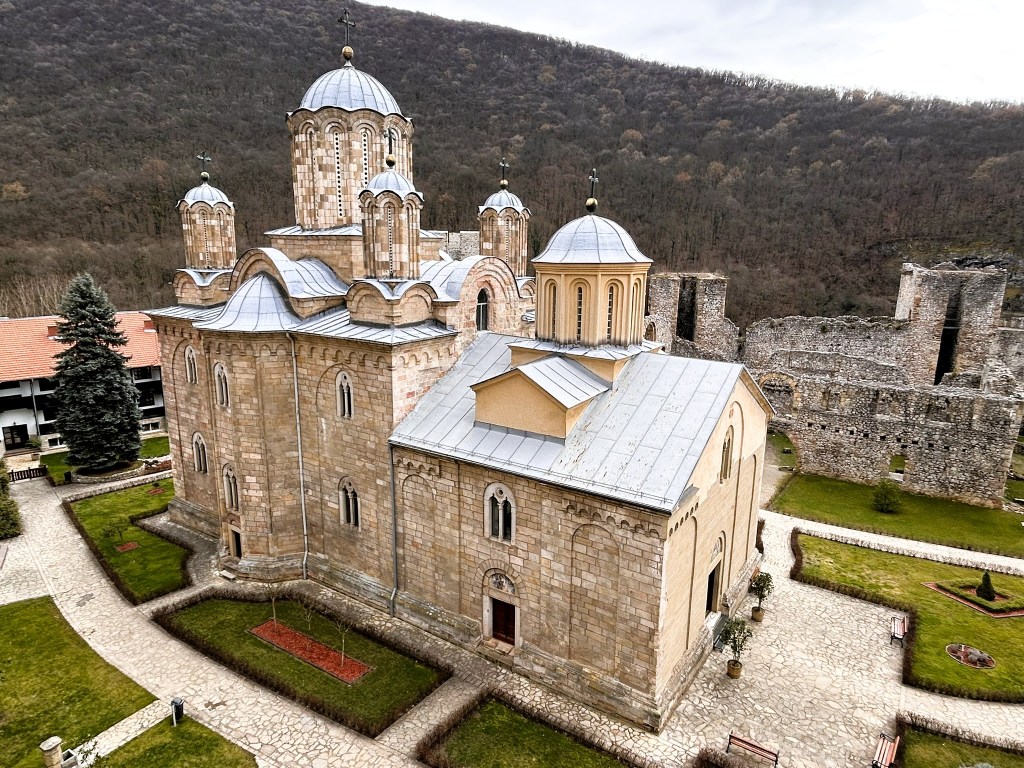 Aerial view of a historic church with domes, surrounded by green gardens and trees, with ancient stone ruins in the background.