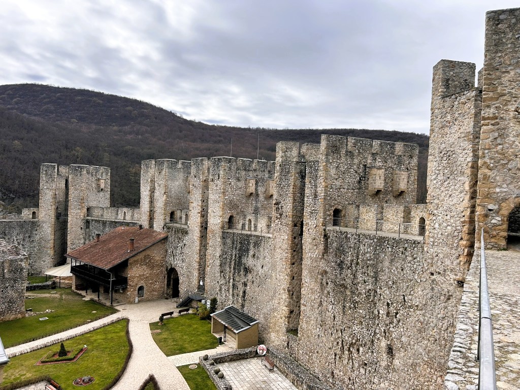 A panoramic view of an ancient stone castle surrounded by a grassy area and trees, under a cloudy sky.