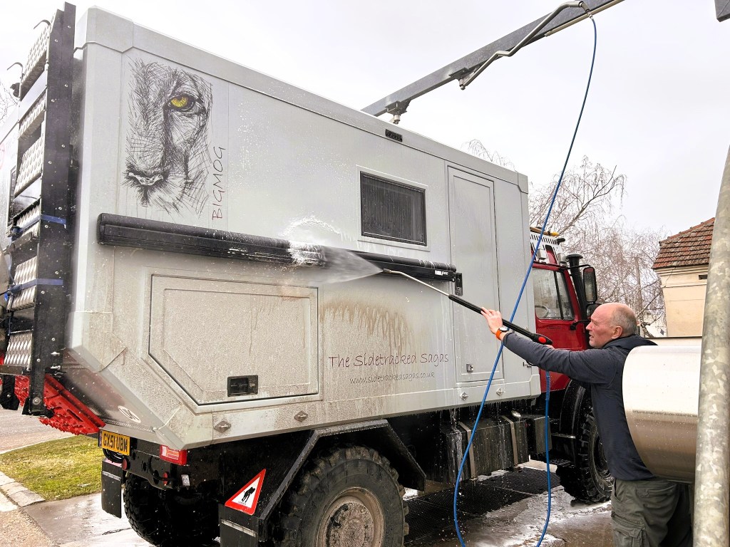 A person washing a large expedition vehicle with a lion design on the side, using a high-pressure water hose.