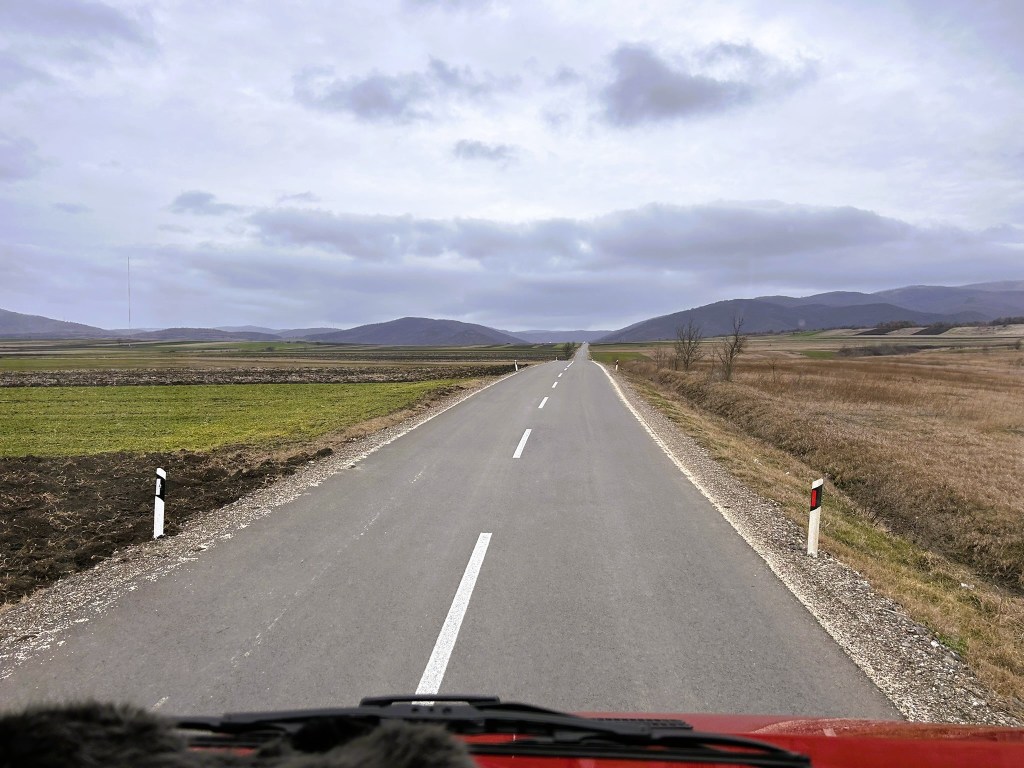 A straight, empty road stretching into the distance, flanked by fields and hills under a cloudy sky.