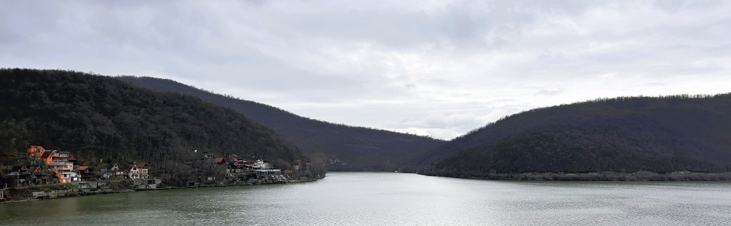 A panoramic view of a calm river surrounded by hills under a cloudy sky, featuring houses along the riverbank.
