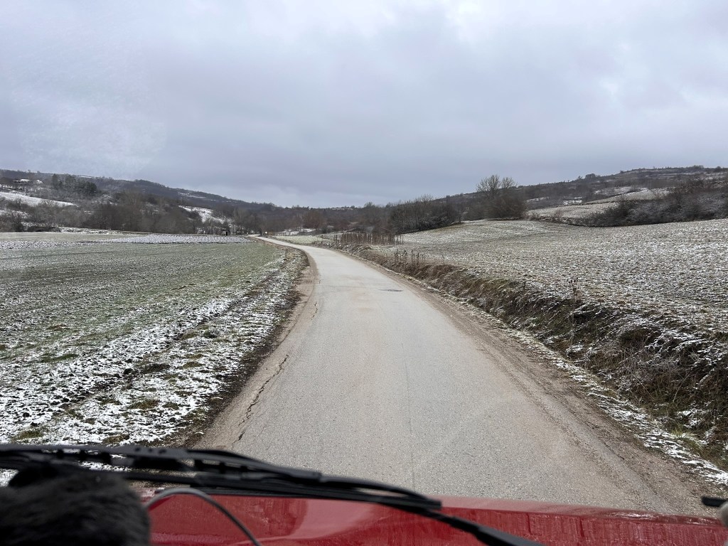 A view from a vehicle driving along a snow-dusted rural road, surrounded by fields and hills under a cloudy sky.