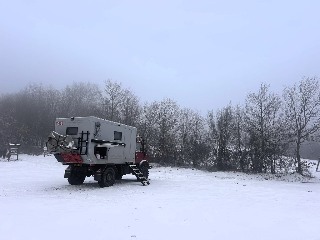 A large expedition vehicle parked in a snowy landscape, surrounded by foggy trees.