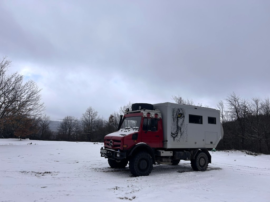 A red expedition vehicle parked on a snowy landscape with trees in the background and cloudy skies.