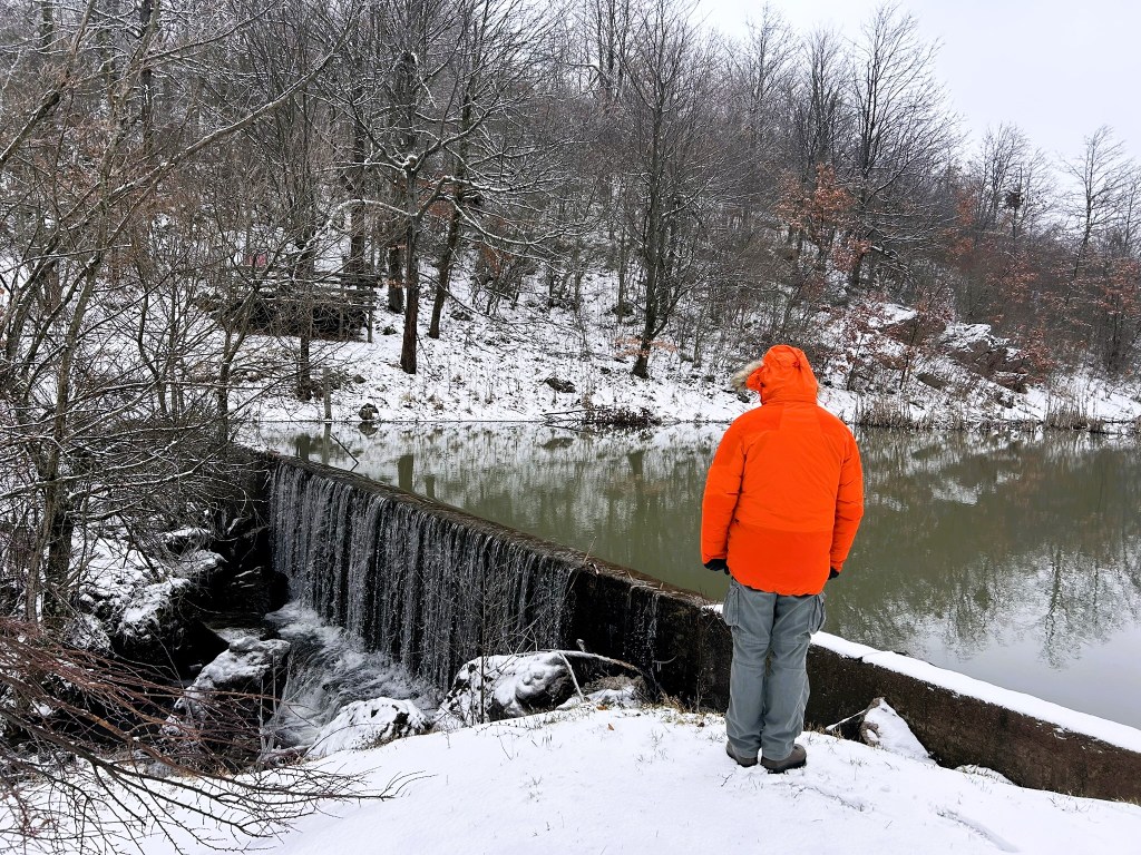 A person in an orange coat stands by a snow-covered riverbank, overlooking a small waterfall and a calm, reflective body of water surrounded by bare trees.