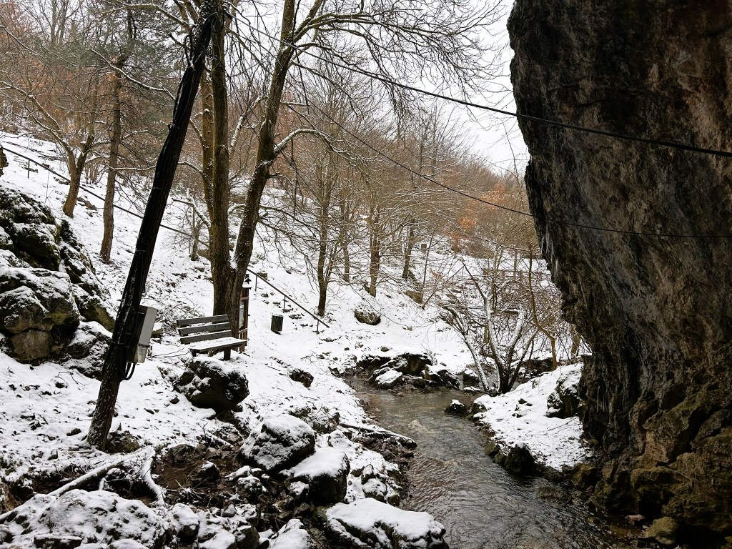 A snowy landscape featuring a creek flowing through a rocky area, with trees in the background and a bench nearby.