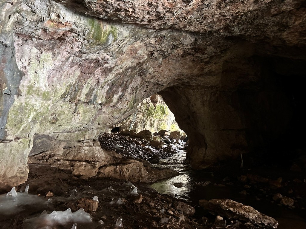 Inside a cave with rocky walls and a stream flowing through, illuminated by light at the entrance.