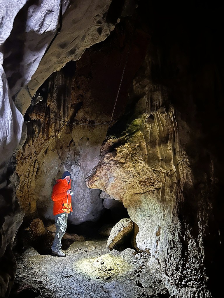A person in a bright orange jacket exploring a cave, illuminated by a flashlight, with rocky walls and uneven terrain.