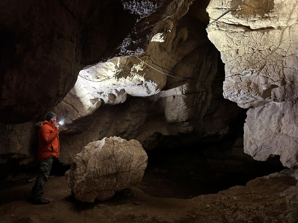 A person wearing an orange jacket stands in a cave, holding a flashlight that illuminates the rocky walls and formations around them.