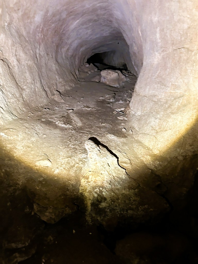 An underground tunnel with rocky walls, dimly lit by a light source illuminating the ground, showing a rough, uneven path and scattered stones.