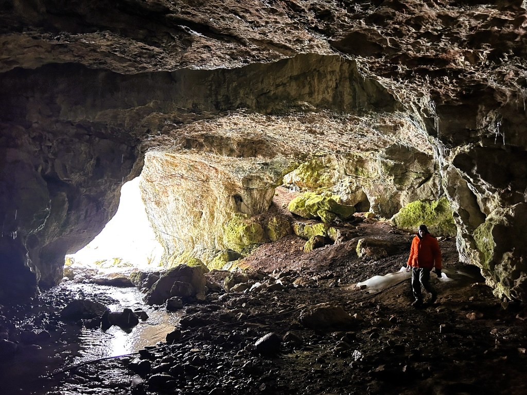 A person in an orange jacket standing inside a large cave, with light illuminating the cave entrance and moss-covered rocks on the ground.