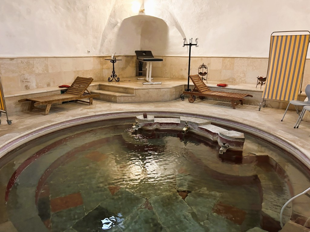 A serene spa environment featuring a circular hot tub with clear water, surrounded by stone walls. Several wooden loungers are positioned nearby, alongside a small table and a coat rack. The lighting is soft, creating a calming atmosphere.