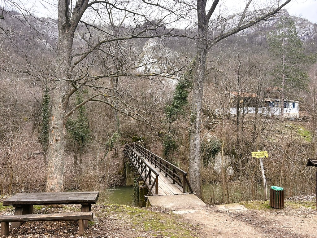 A wooden bridge crosses a small stream surrounded by bare trees and rocky hills. In the background, there is a modest house and a sign, while a picnic table and bin are visible in the foreground.