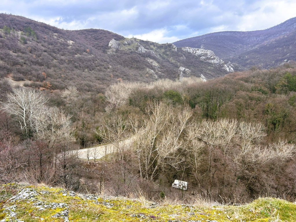 Scenic view of a forested valley with bare trees and rocky hills under a cloudy sky, featuring a winding road and a caravan in the distance.