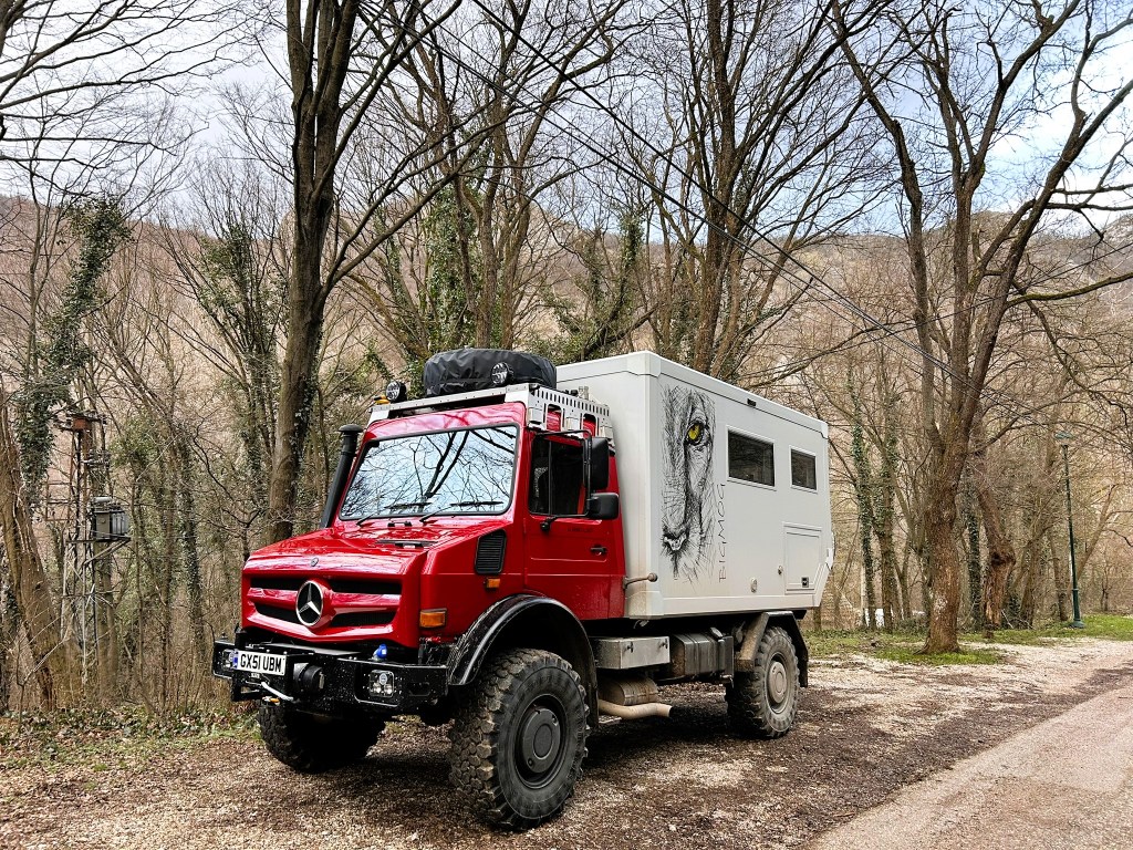 A red Mercedes off-road vehicle with a grey camper shell, adorned with a lion graphic, parked on a gravel path surrounded by bare trees and hills.