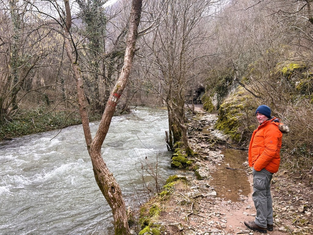 A man in an orange jacket and a blue beanie stands by a rushing river, surrounded by bare trees and a rocky path in a natural setting.
