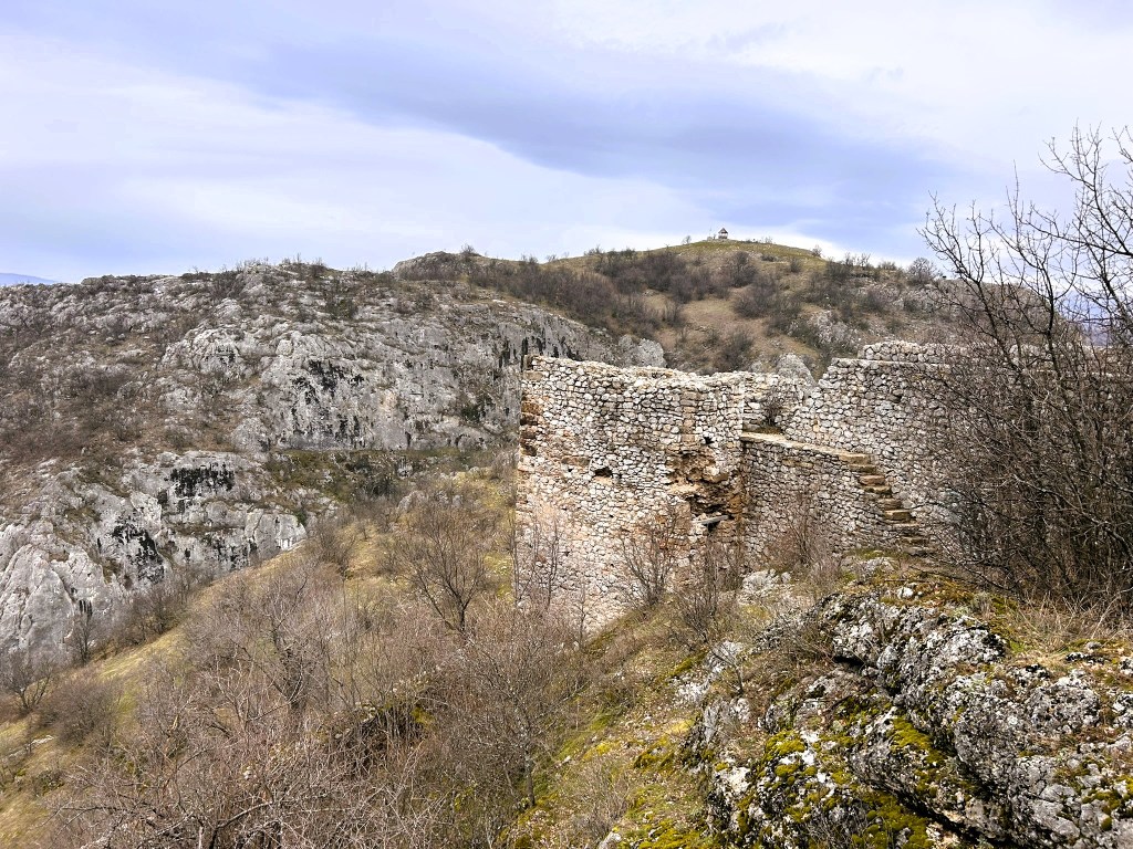 Ruins of an old stone structure on a hillside with rocky terrain and sparse vegetation under a cloudy sky.