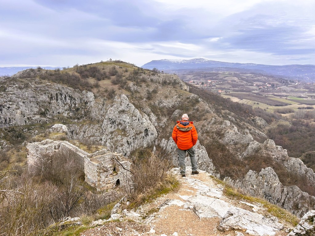 A person in an orange jacket stands on a rocky ledge, overlooking a hilly landscape with ruins and mountains in the distance under a cloudy sky.