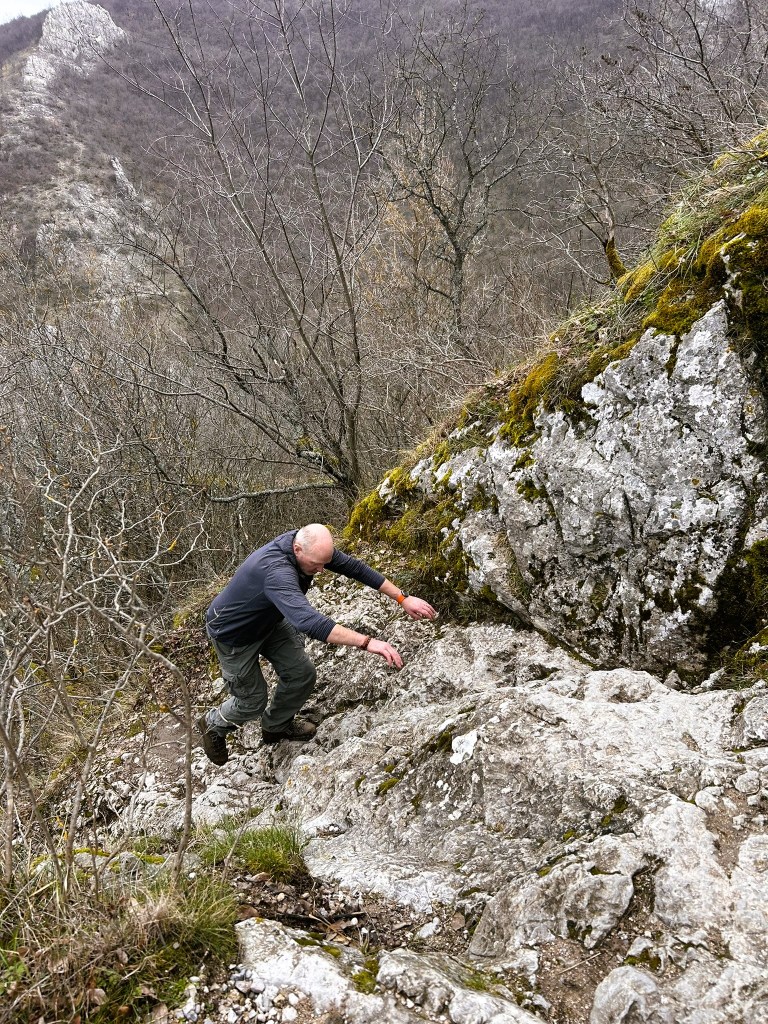 A man climbing a rocky path in a mountainous area, surrounded by bare trees and rocky terrain.