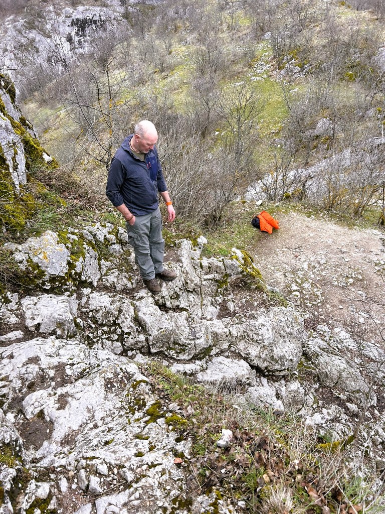 A person stands on rocky terrain, gazing downwards, surrounded by sparse trees and a hilly landscape.