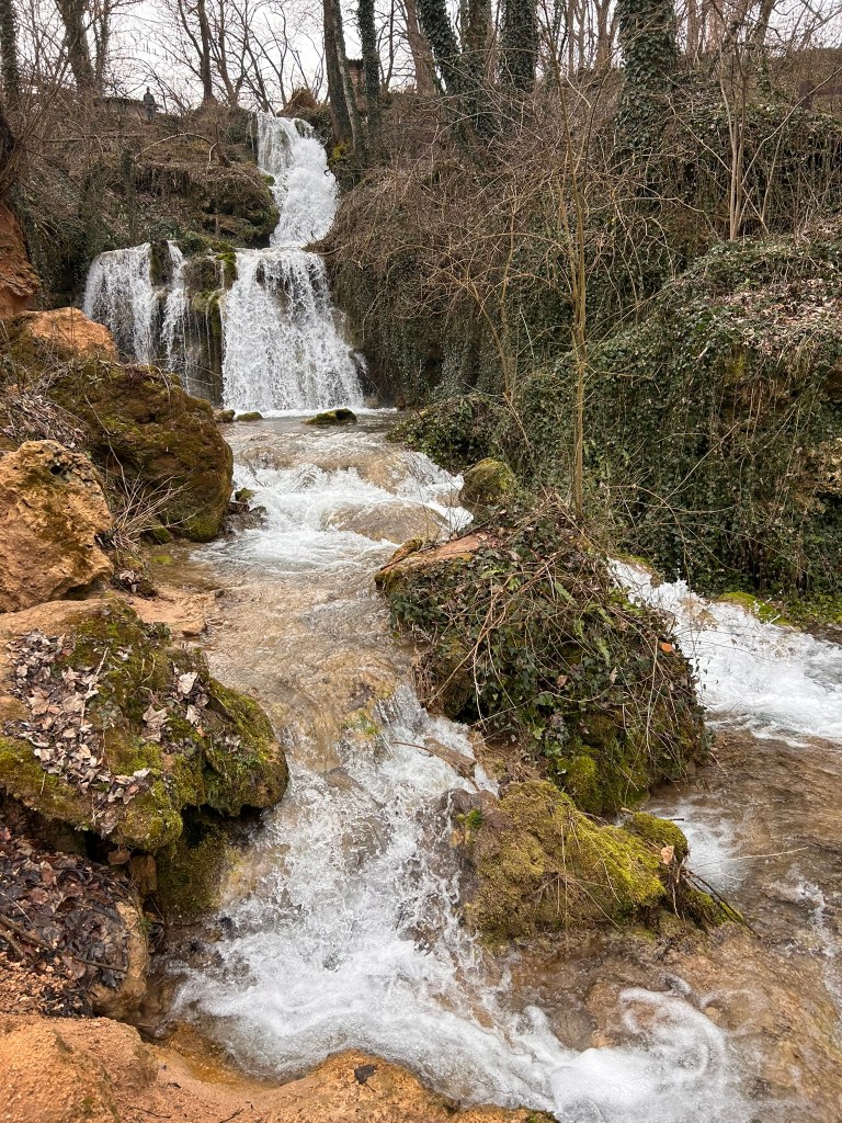 A serene landscape featuring a cascading waterfall surrounded by rocks and lush greenery, with water flowing gently over a rocky stream.