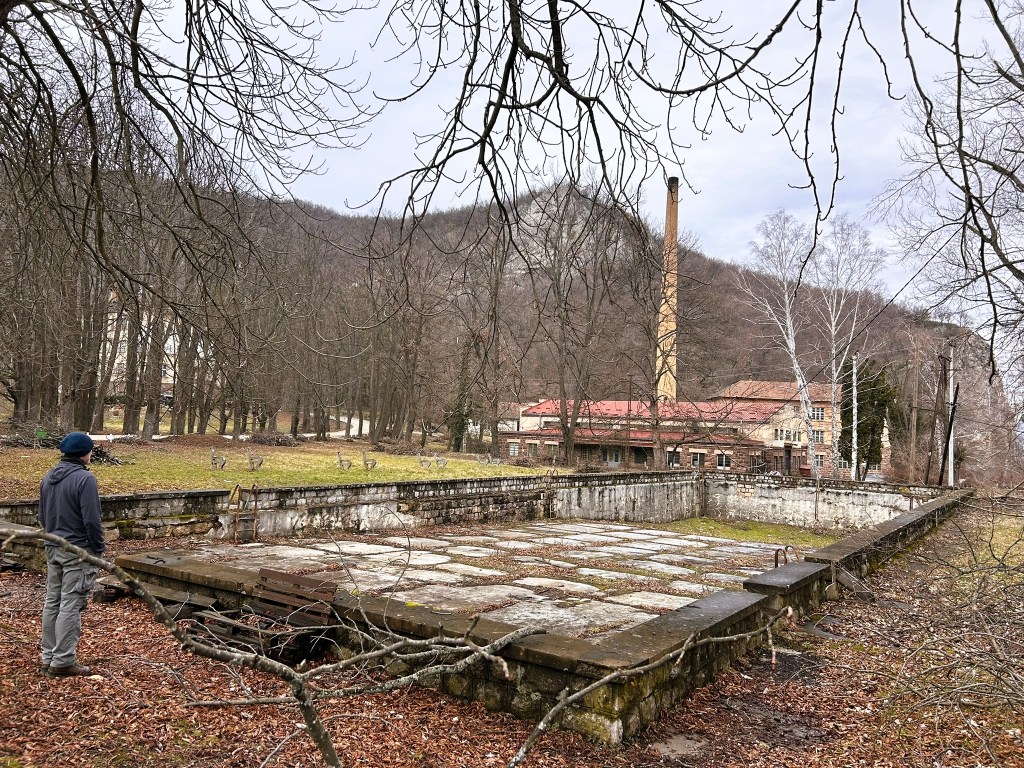 A person stands on a stone platform in an overgrown area, with an abandoned building and chimney visible in the background, surrounded by leafless trees and mountains.