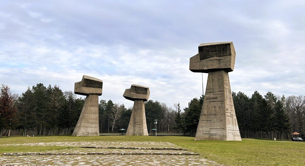 A landscape featuring three large, abstract concrete monuments on grassy ground, surrounded by trees under a cloudy sky.