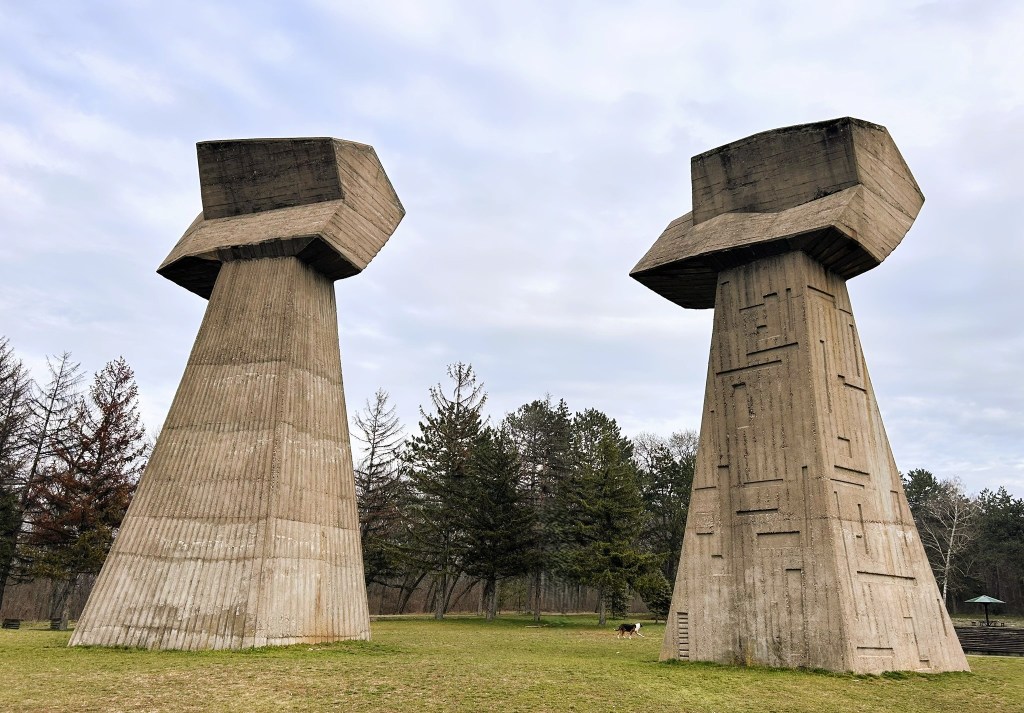 Two large, abstract concrete sculptures resembling towers in a grassy landscape, surrounded by trees under a cloudy sky.