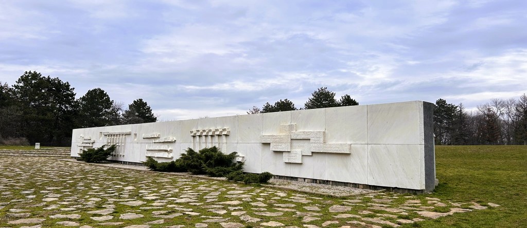 A modern stone monument with abstract carvings, set on a grassy area surrounded by trees under a cloudy sky.