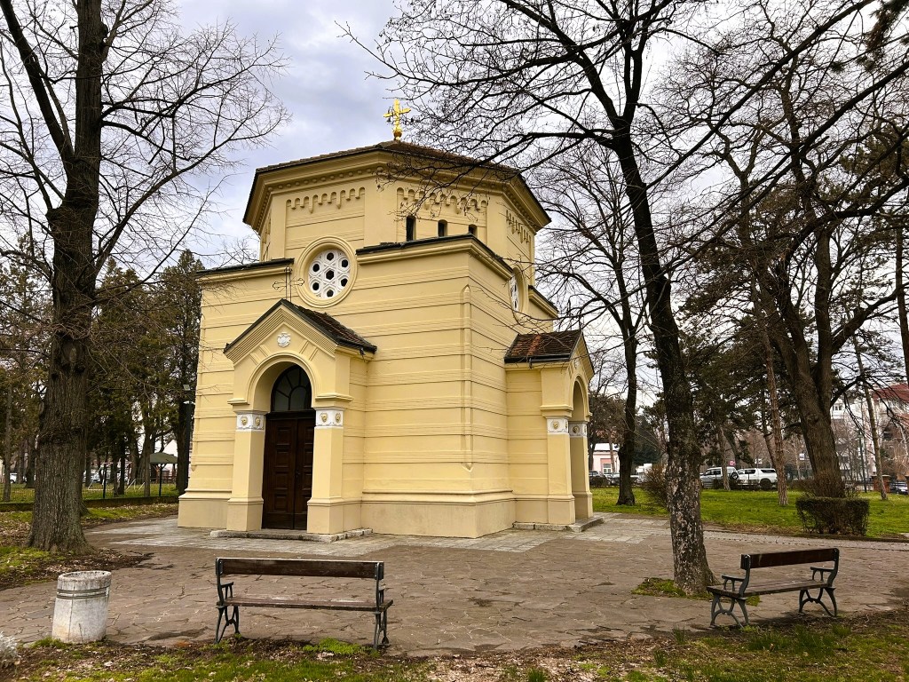A small yellow church with architectural details, surrounded by trees in a park setting, featuring benches and a pathway.