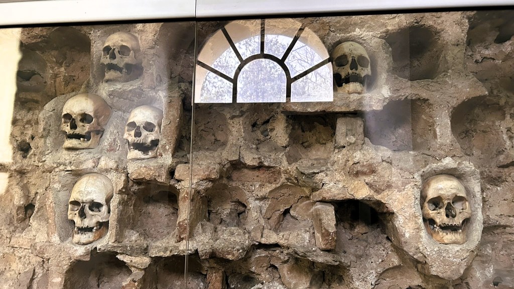 A wall displaying several human skulls set in stone niches, with a small arched window in the upper section.