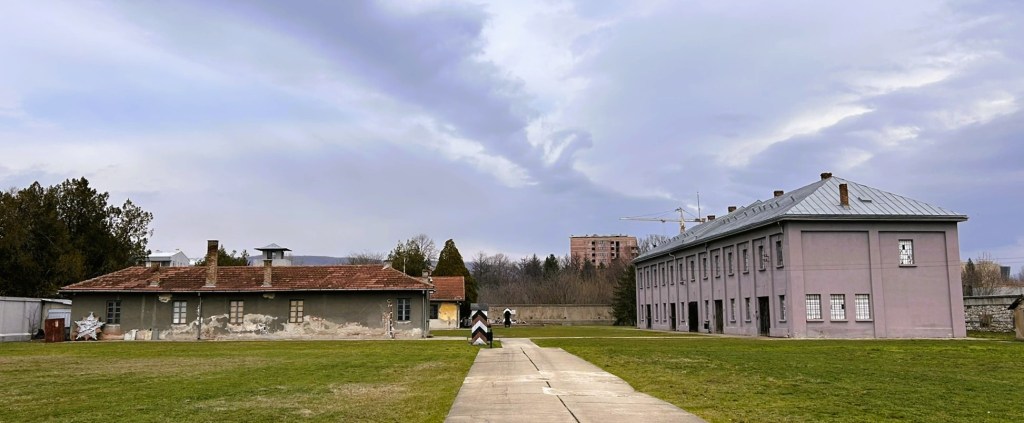 A historical site featuring two buildings, one with a red tiled roof and the other in grey with multiple windows, set against a cloudy sky and green lawn.