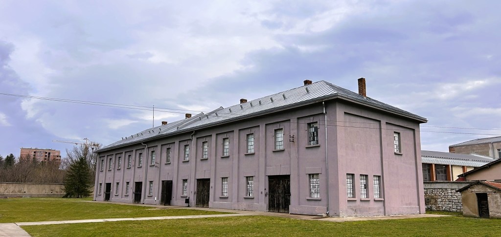Exterior view of a grey building with a pitched roof, featuring barred windows and grassy surroundings under a cloudy sky.
