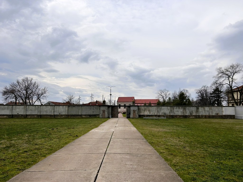 A wide pathway leading towards a gate in a concrete wall, flanked by grass and trees under a cloudy sky.