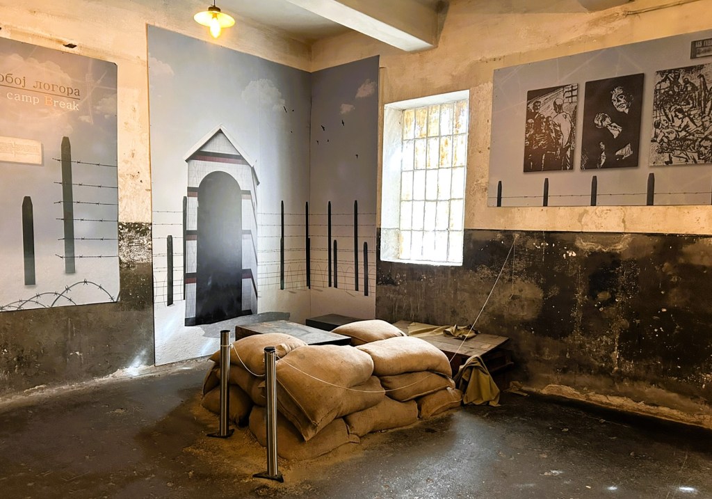 Interior of a historical exhibition space featuring a mural of a prison camp entrance, barbed wire, and photographs depicting camp life, with sandbags and barriers in the foreground.