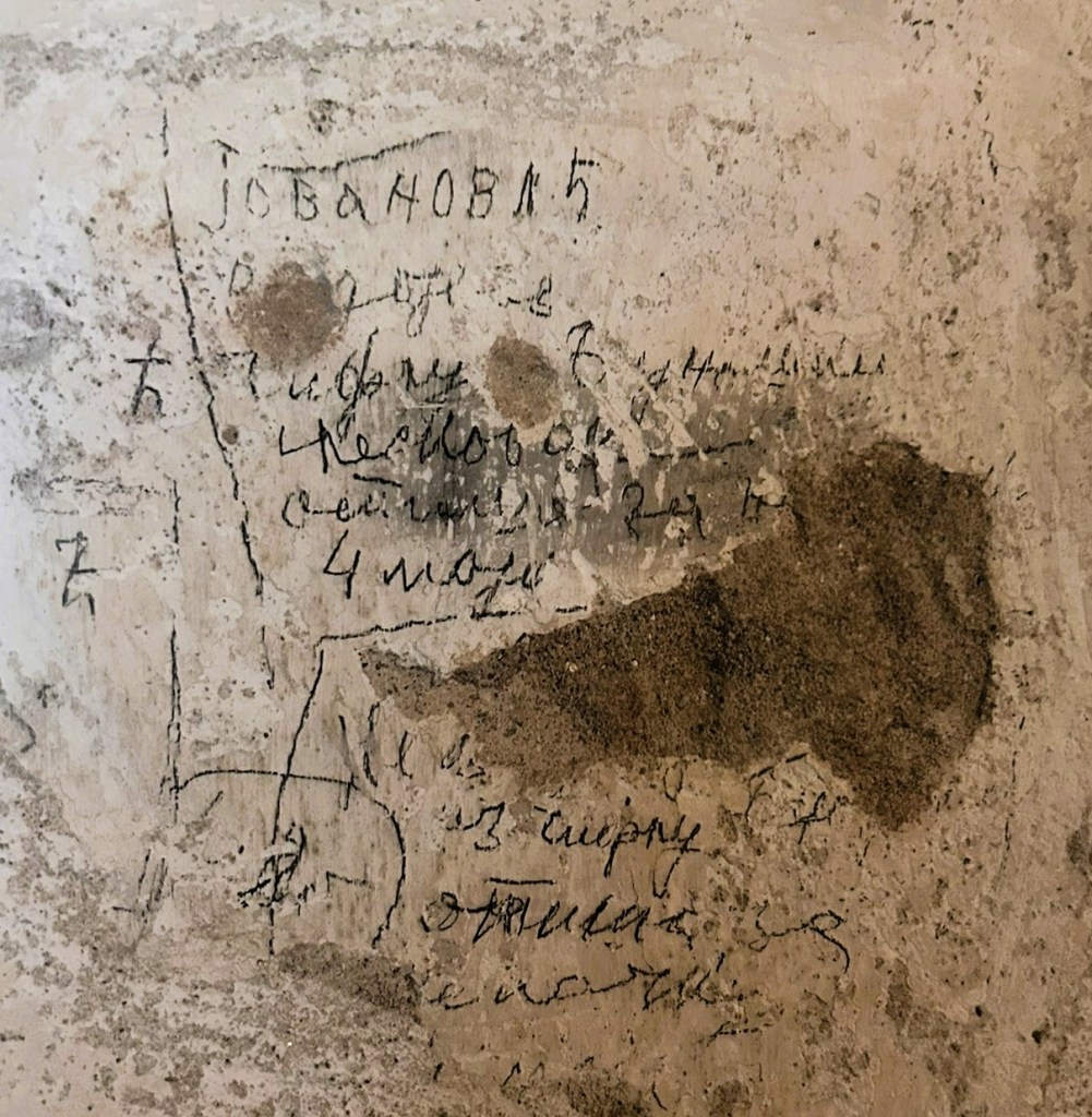 A textured wall featuring handwritten inscriptions and markings, partially obscured by dust and dirt.