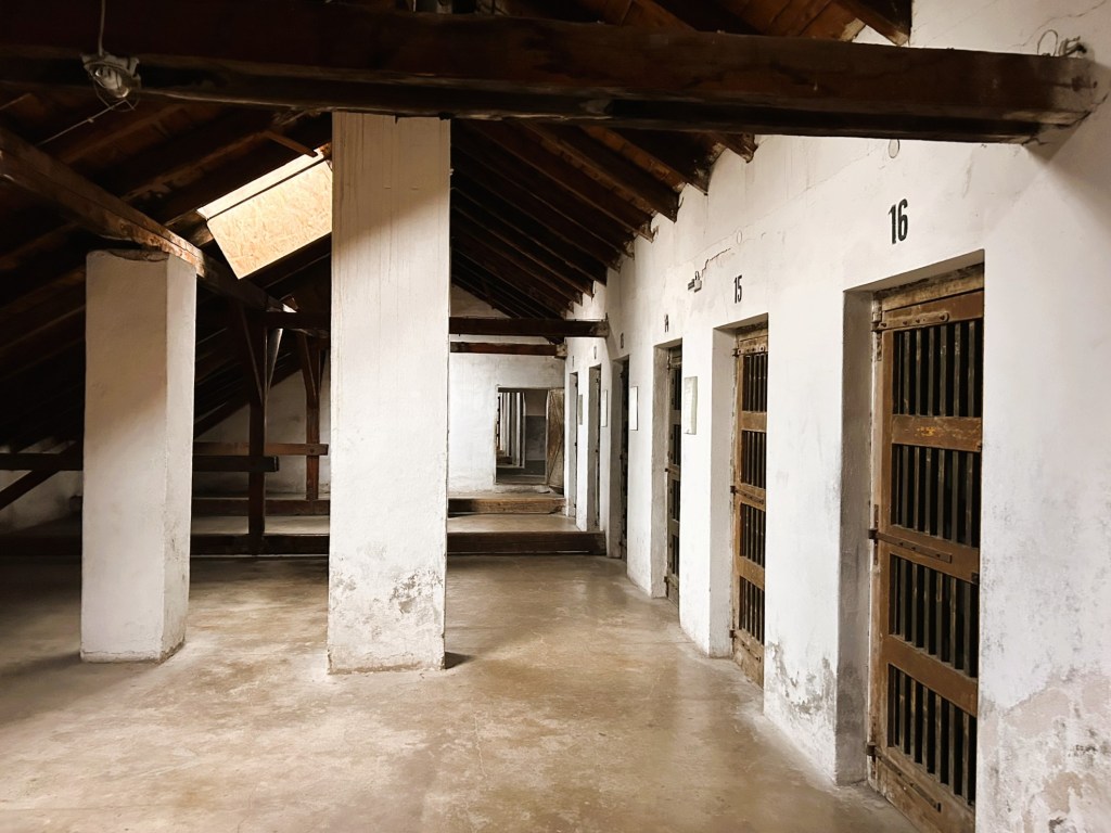 Interior of an old prison building featuring multiple jail cells with wooden doors and barred windows, under a slanted wooden roof.