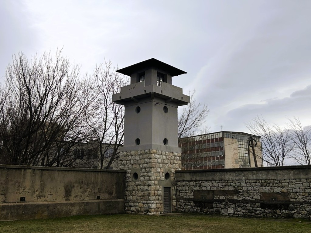A modern observation tower with a flat roof, situated next to a stone wall and surrounded by leafless trees, set against a cloudy sky.