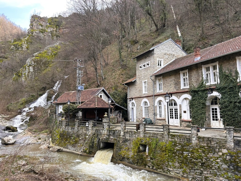 A scenic view of a small stone house next to a river, with a waterfall flowing nearby. The background features a rocky hillside and bare trees, creating a tranquil natural setting.